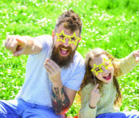 Child and dad posing with star shaped eyeglases photo booth attribute at meadow. Father and daughter sits on grass at free Reno concerts.