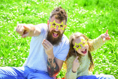 Child and dad posing with star shaped eyeglases photo booth attribute at meadow. Father and daughter sits on grass at free Reno concerts.