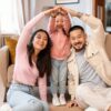 Happy family forming a house shape with their arms while sitting on the living room floor, symbolizing the concept of achieving the dream of homeownership.