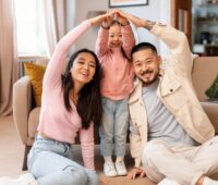 Happy family forming a house shape with their arms while sitting on the living room floor, symbolizing the concept of achieving the dream of homeownership.