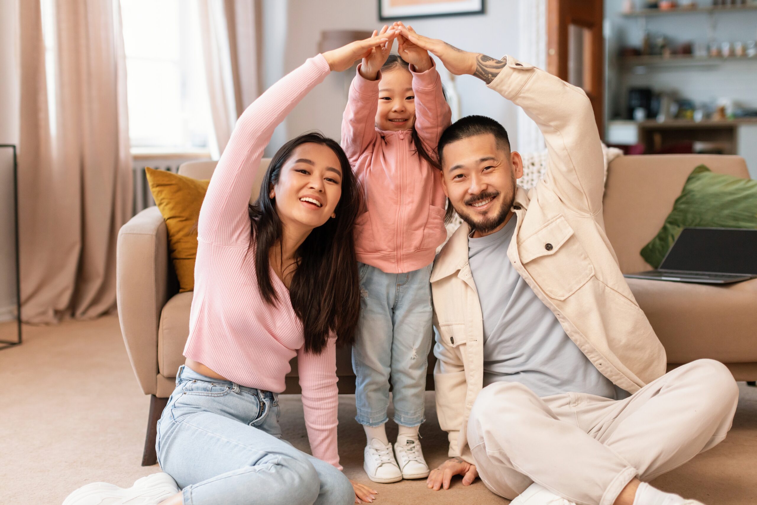 Happy family forming a house shape with their arms while sitting on the living room floor, symbolizing the concept of achieving the dream of homeownership.