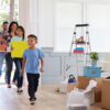 A family of four enters their new home, smiling and carrying moving boxes. A young boy leads the way, followed by his older sister and parents. The room is filled with unpacked boxes, a ladder, and home decor items, creating a sense of excitement and new beginnings.