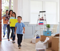 A family of four enters their new home, smiling and carrying moving boxes. A young boy leads the way, followed by his older sister and parents. The room is filled with unpacked boxes, a ladder, and home decor items, creating a sense of excitement and new beginnings.