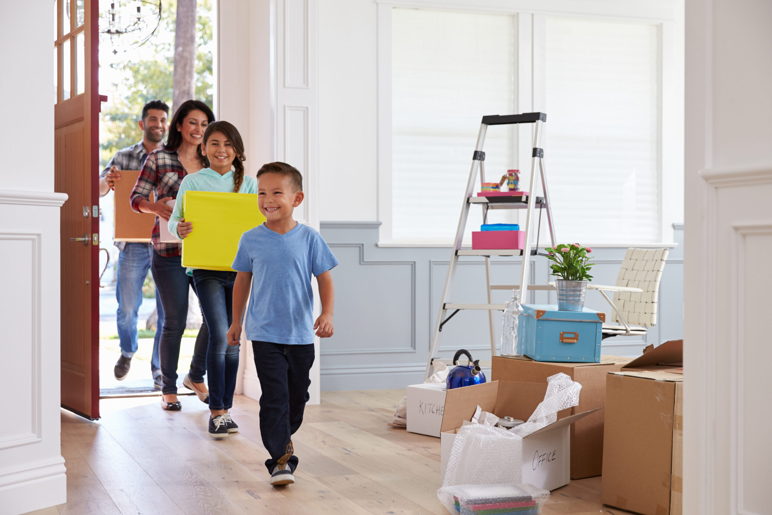 A family of four enters their new home, smiling and carrying moving boxes. A young boy leads the way, followed by his older sister and parents. The room is filled with unpacked boxes, a ladder, and home decor items, creating a sense of excitement and new beginnings.