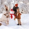 Happy parents and children gathering in snow-covered park together sculpting funny snowman from snow. Father, mother and two kids playing outdoor in winter forest. Family active holiday tradition.