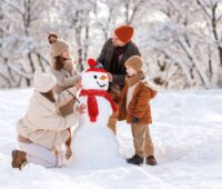 Happy parents and children gathering in snow-covered park together sculpting funny snowman from snow. Father, mother and two kids playing outdoor in winter forest. Family active holiday tradition.