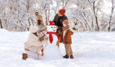 Happy parents and children gathering in snow-covered park together sculpting funny snowman from snow. Father, mother and two kids playing outdoor in winter forest. Family active holiday tradition.