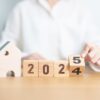 A person in a white shirt is adjusting wooden blocks displaying the numbers 2024 and 2025, with a small wooden house figure positioned to the left, representing housing market predictions for the years ahead.