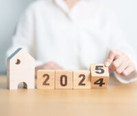 A person in a white shirt is adjusting wooden blocks displaying the numbers 2024 and 2025, with a small wooden house figure positioned to the left, representing housing market predictions for the years ahead.