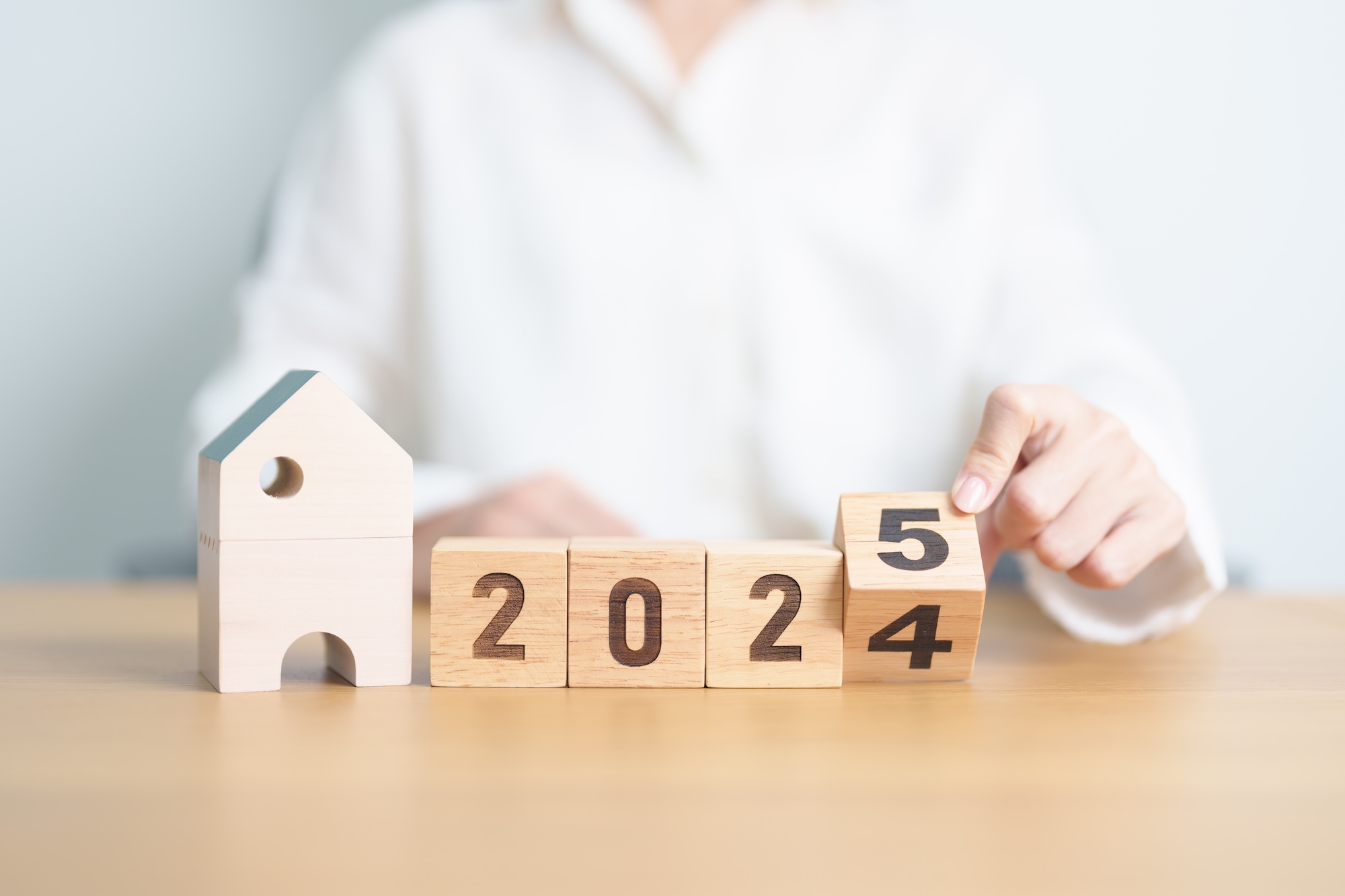 A person in a white shirt is adjusting wooden blocks displaying the numbers 2024 and 2025, with a small wooden house figure positioned to the left, representing housing market predictions for the years ahead.