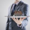 A luxury real estate agent in a suit elegantly presents a silver tray, showcasing a small model house with sophistication and charm.