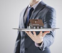 A luxury real estate agent in a suit elegantly presents a silver tray, showcasing a small model house with sophistication and charm.