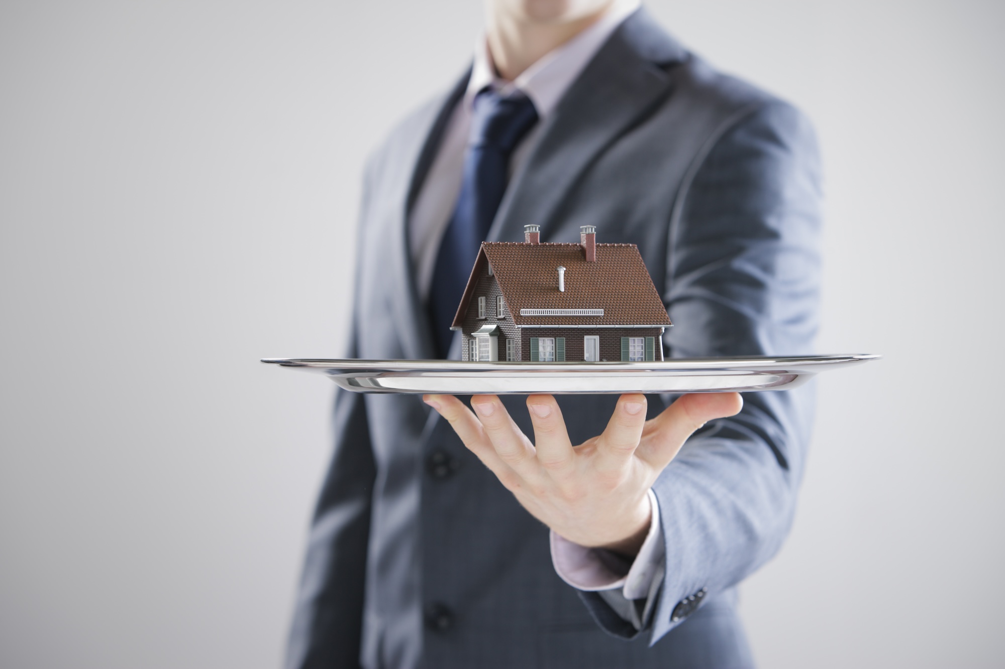 A luxury real estate agent in a suit elegantly presents a silver tray, showcasing a small model house with sophistication and charm.